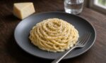 Koyu gri tabakta kremamsı pecorino sosuna bulanmış spagetti, üzerinde iri çekilmiş karabiber; yanında çatal, bir parça pecorino ve bir bardak su