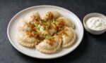 Koyu arduvaz masada kırık beyaz taş tabakta kenarları kıvrımlı yarım ay pierogiler, üzerlerinde tereyağında karamelize soğan ve doğranmış frenk soğanı, yanında kâsede ekşi krema
